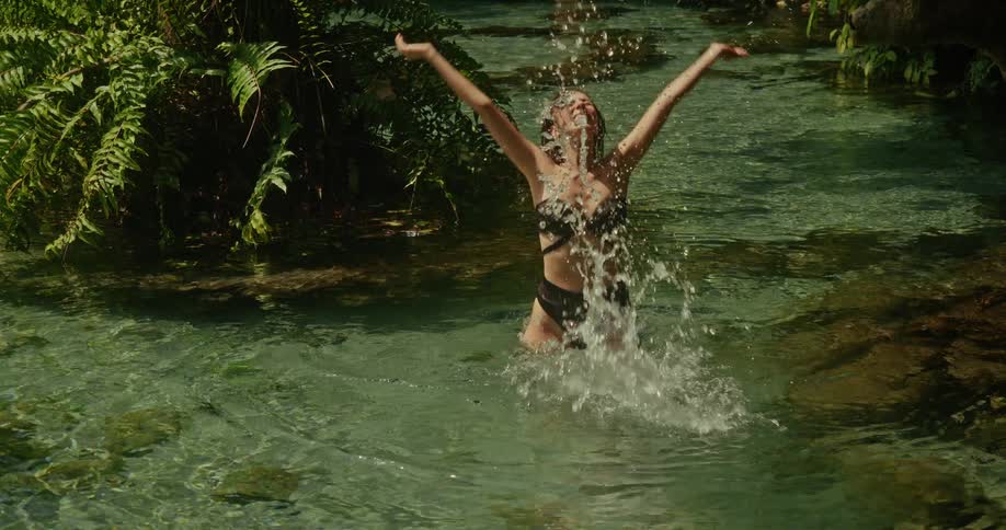 A beautiful woman in a black bikini joyfully splashes in a pristine, crystal-clear jungle stream in slow motion
