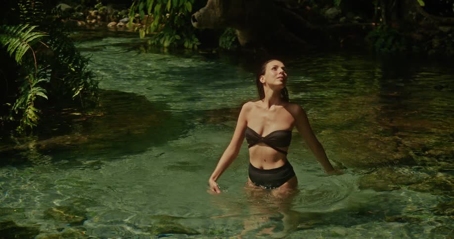 A graceful woman in a black bikini enjoys the serene atmosphere while standing in the clear, cool waters of a secluded jungle stream