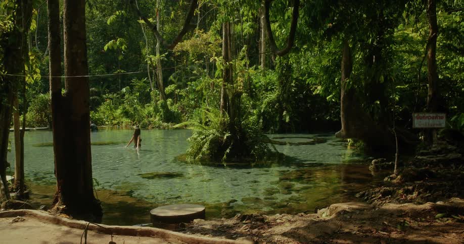 A beautiful woman in a bikini wades through a crystal clear jungle spring surrounded by lush tropical greenery