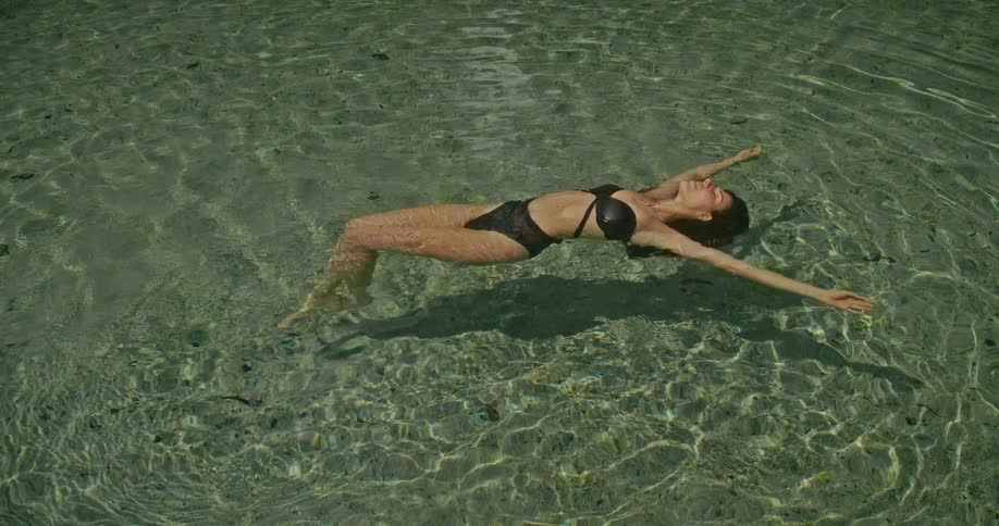 A serene woman in a black swimsuit floats peacefully on her back in the crystal-clear waters of a tranquil jungle spring