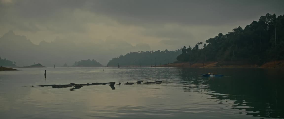 A lone kayaker paddles across a serene and misty lake surrounded by lush green mountains on an overcast day
