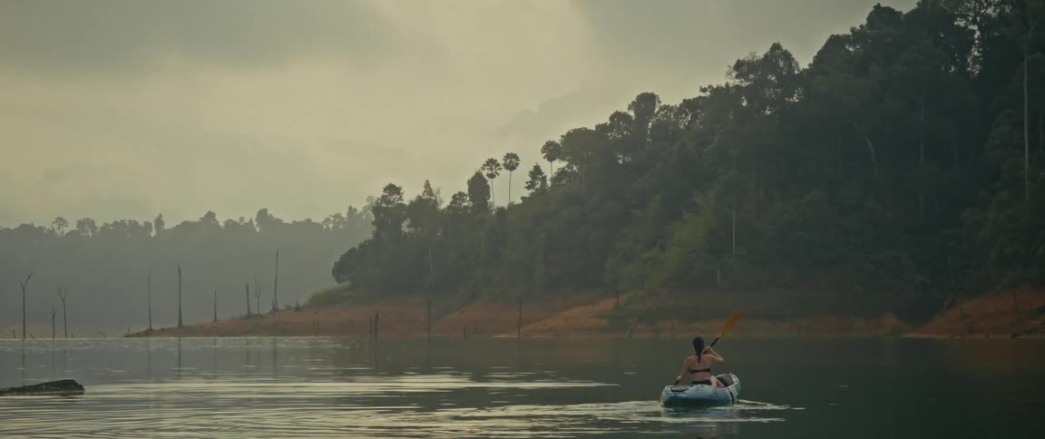 An adventurous woman enjoys a peaceful journey while kayaking alone on a serene, misty lake surrounded by a lush tropical jungle