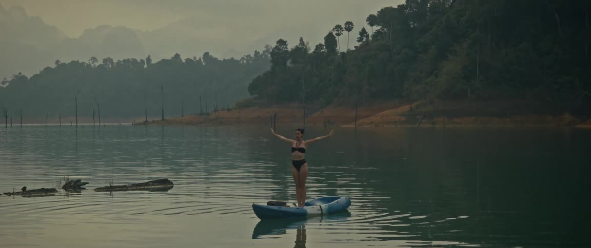 A woman in a bikini finds her balance practicing a yoga pose while standing on a kayak in a tranquil, misty lake