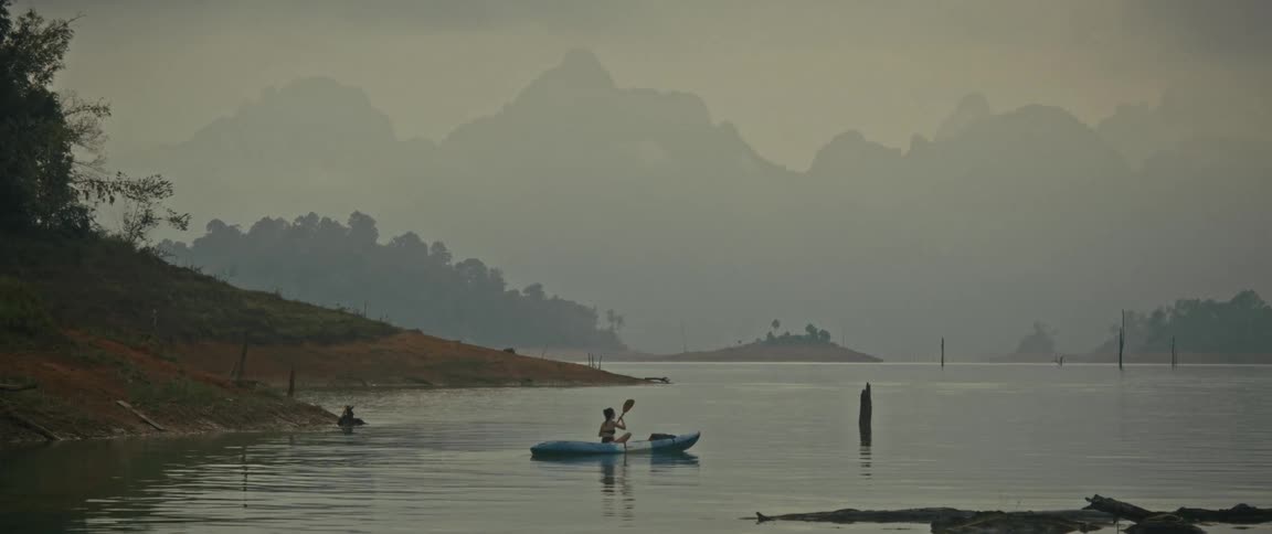 A lone kayaker peacefully paddles across a serene, misty lake surrounded by majestic mountains at dawn