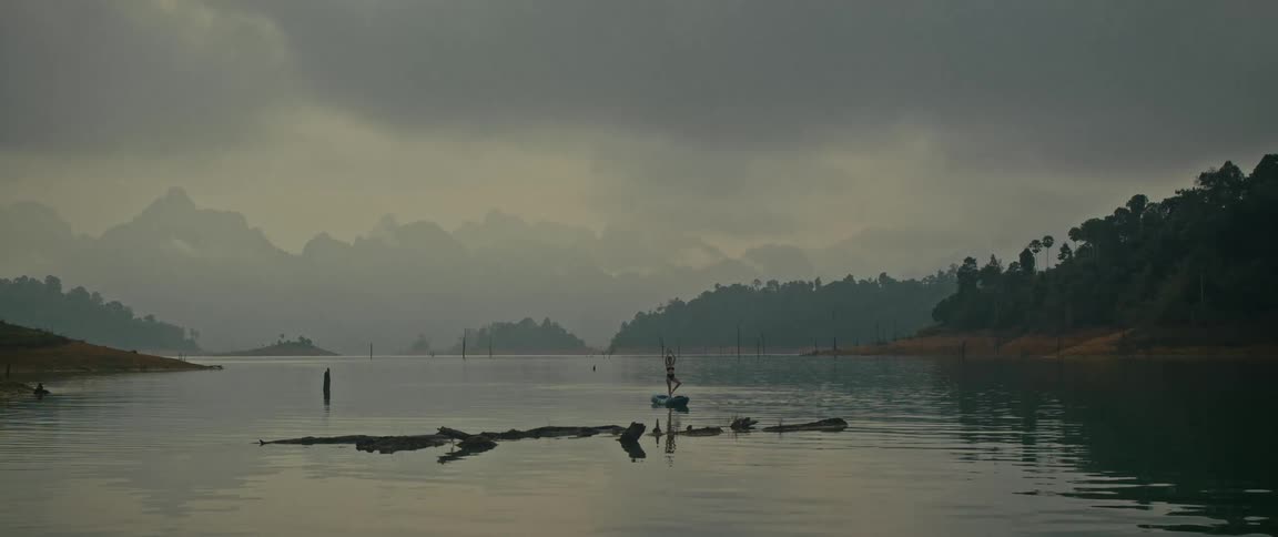 A solitary person practices a yoga pose on a stand-up paddleboard on a vast, tranquil lake surrounded by misty mountains