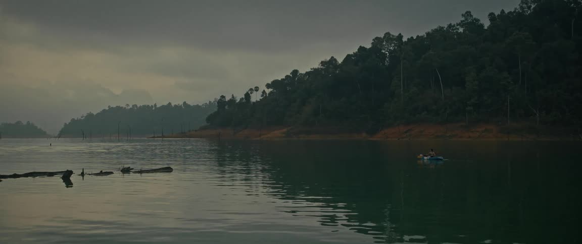 Two people in a kayak paddle across a tranquil lake surrounded by a lush forest on a cloudy day