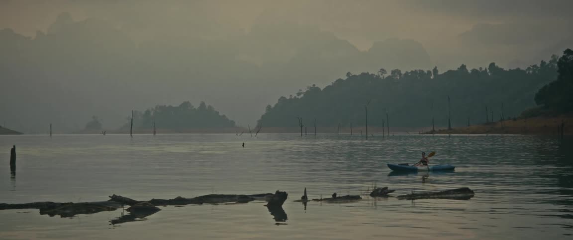 A person paddles a kayak across a calm, misty lake with a vast, mountainous, and forested landscape in the background