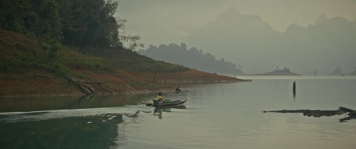 A local in a motorboat glides across a tranquil, misty lake as a woman kayaks in the foreground, surrounded by hazy, jungle-clad mountains