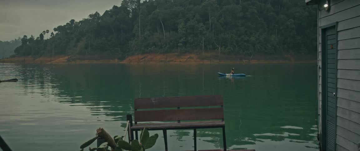 A slow zoom shot captures a serene view of a person kayaking on a tranquil green lake surrounded by a lush, dense forest