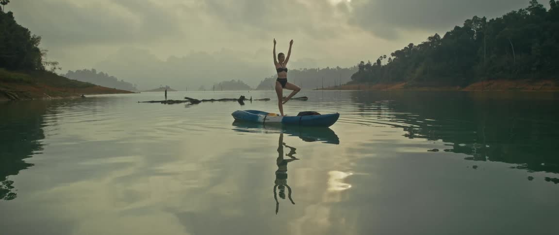 A serene woman in a bikini practices a yoga tree pose while balancing on a kayak in the middle of a tranquil lake surrounded by misty mountains