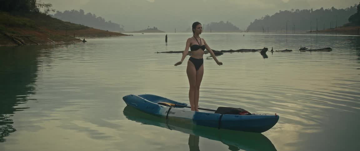 A woman in a black bikini practices yoga on a kayak, peacefully floating on a calm lake surrounded by misty mountains