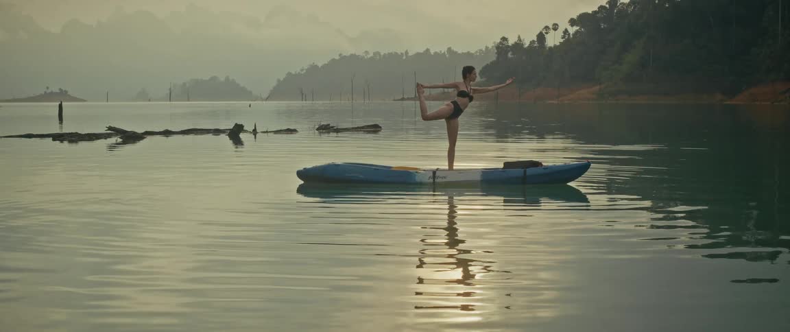 A woman gracefully performs a standing bow yoga pose on a kayak, floating on a tranquil, misty lake