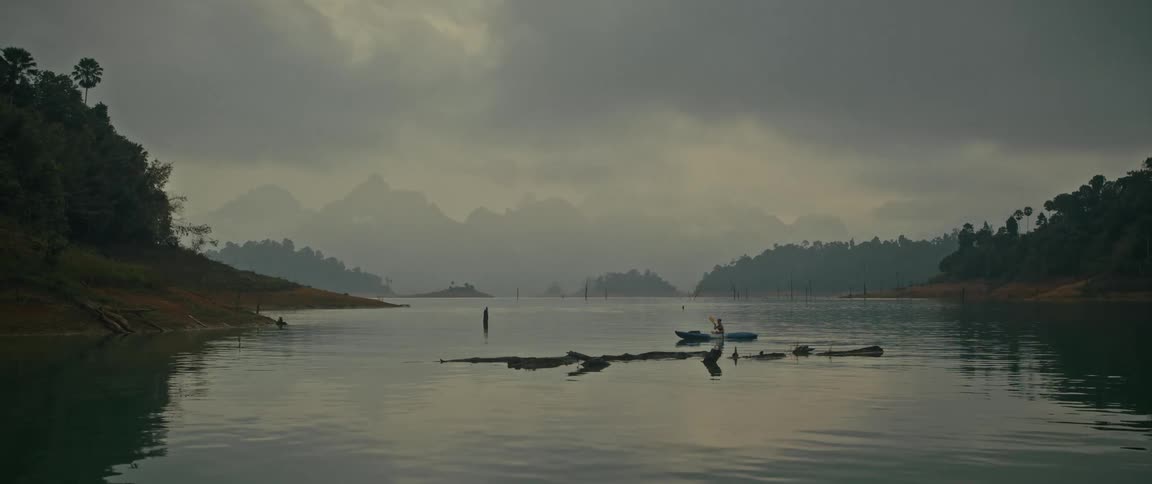 A lone kayaker paddles across a serene lake surrounded by misty mountains on a cloudy day