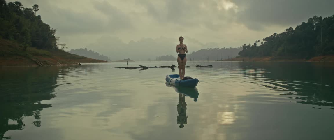 A woman in a bikini meditates on a blue kayak in the middle of a vast, calm lake surrounded by mountains