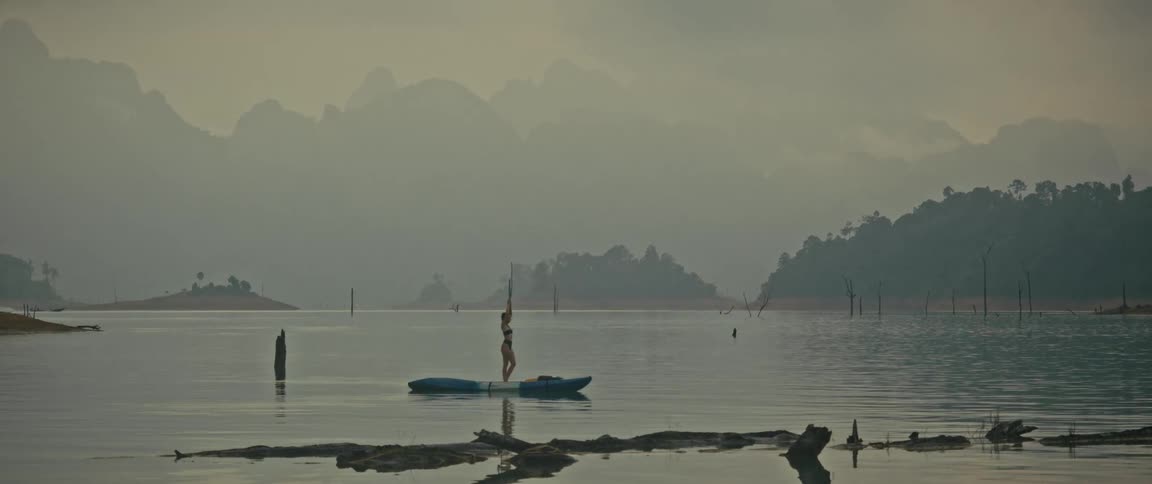 A serene woman in a bikini practices yoga poses while balancing on a kayak in the middle of a calm, misty lake surrounded by mountains