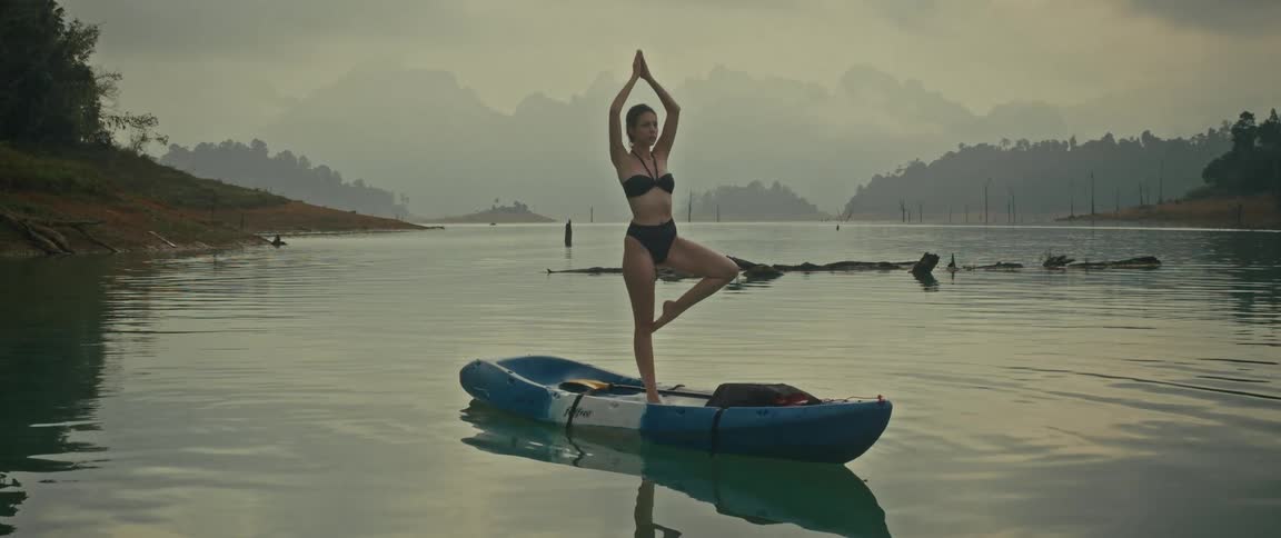 A woman in a bikini practices yoga, balancing in a tree pose on a kayak floating in a serene mountain lake