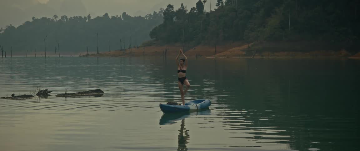 A woman in a bikini finds her balance and practices a yoga tree pose while standing on a kayak in the middle of a tranquil, misty lake