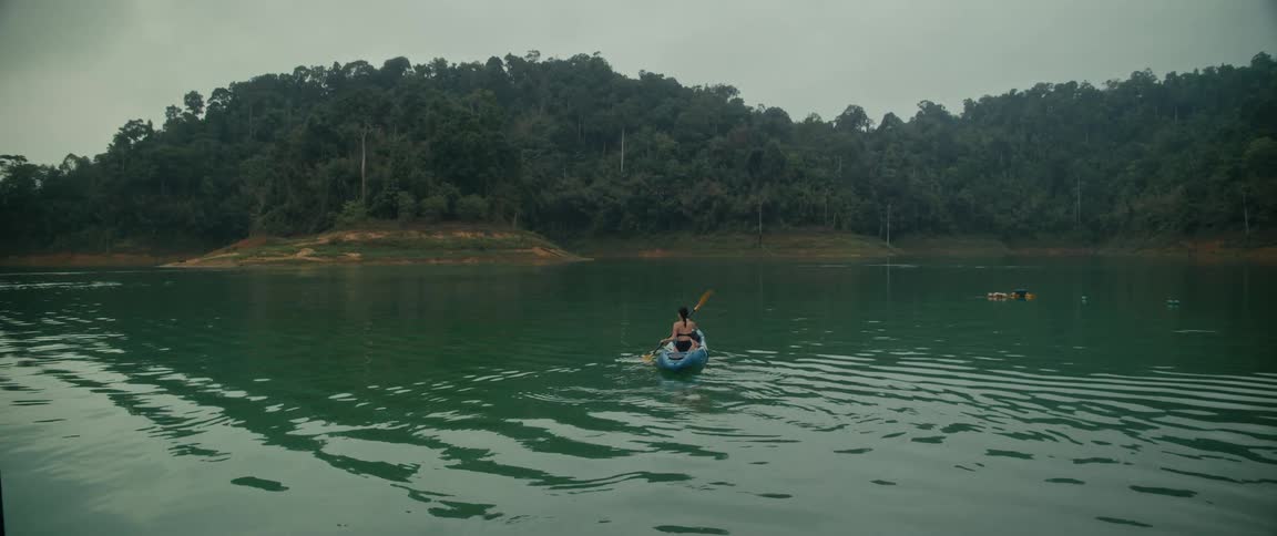 A woman kayaks on a tranquil green lake surrounded by a dense forest, with the camera slowly pulling back to reveal the view from a peaceful lakeside bungalow