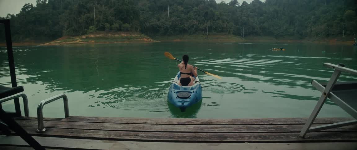 Slow-motion view from a lakeside cabin of a woman in a bikini paddling a kayak across a tranquil green lake surrounded by a forest