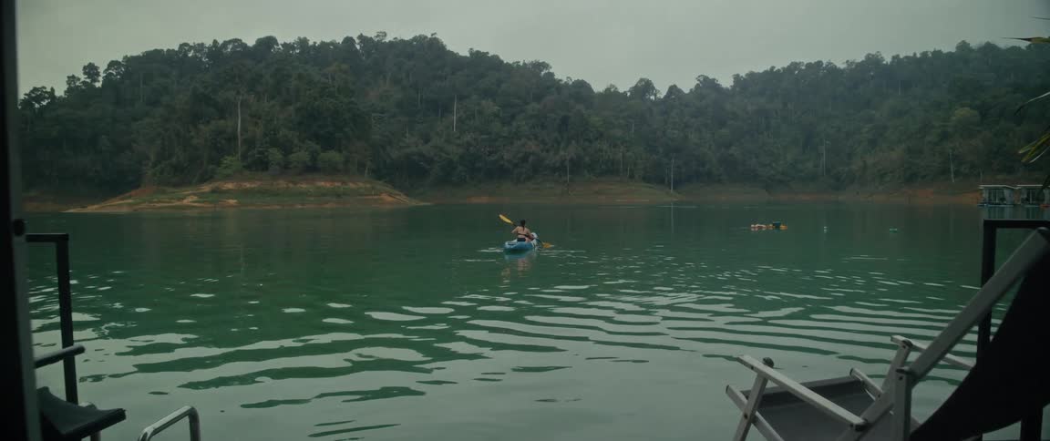 Slow zoom from a cabin porch onto a person kayaking on a tranquil green lake surrounded by a lush forest