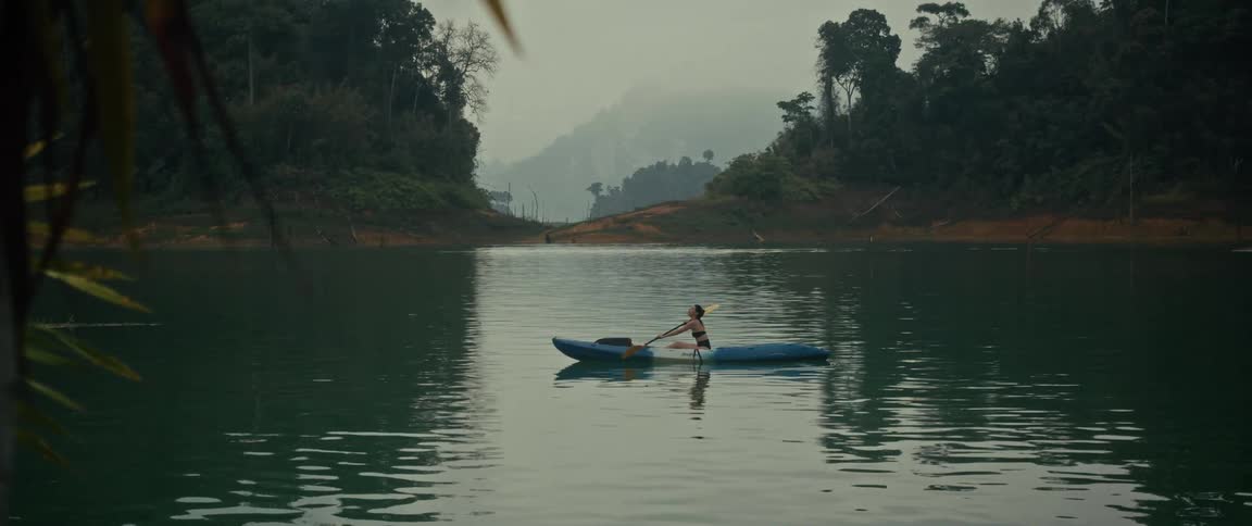 A lone kayaker peacefully paddles in slow motion across a serene, misty lake surrounded by dense jungle hills