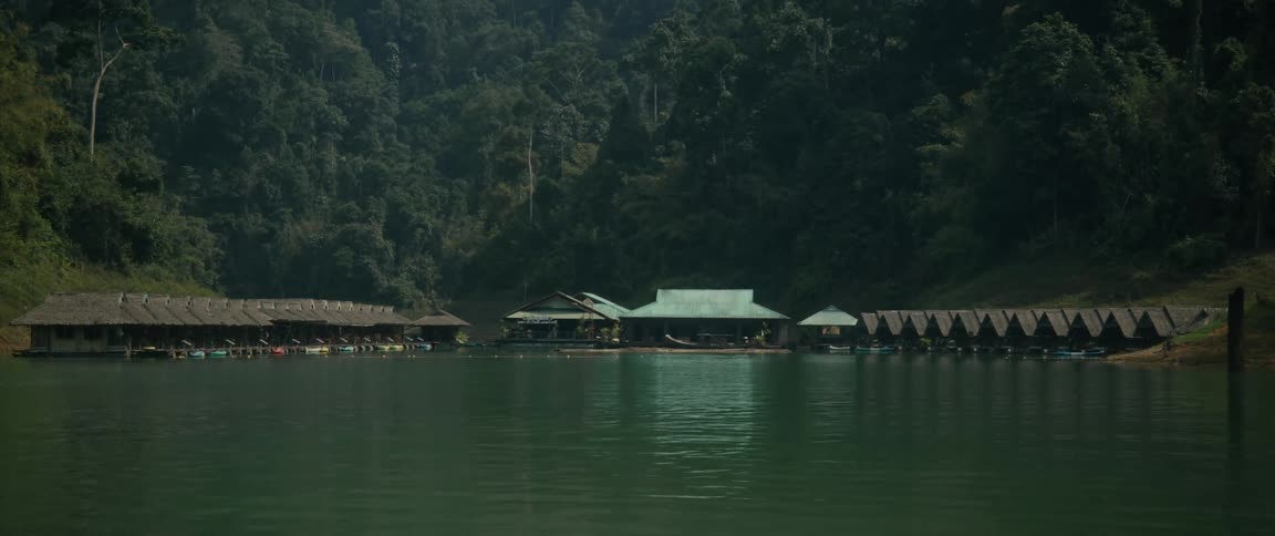 A scenic wide shot of a peaceful floating bungalow resort on a tranquil green lake, nestled at the foot of a lush, forested mountain