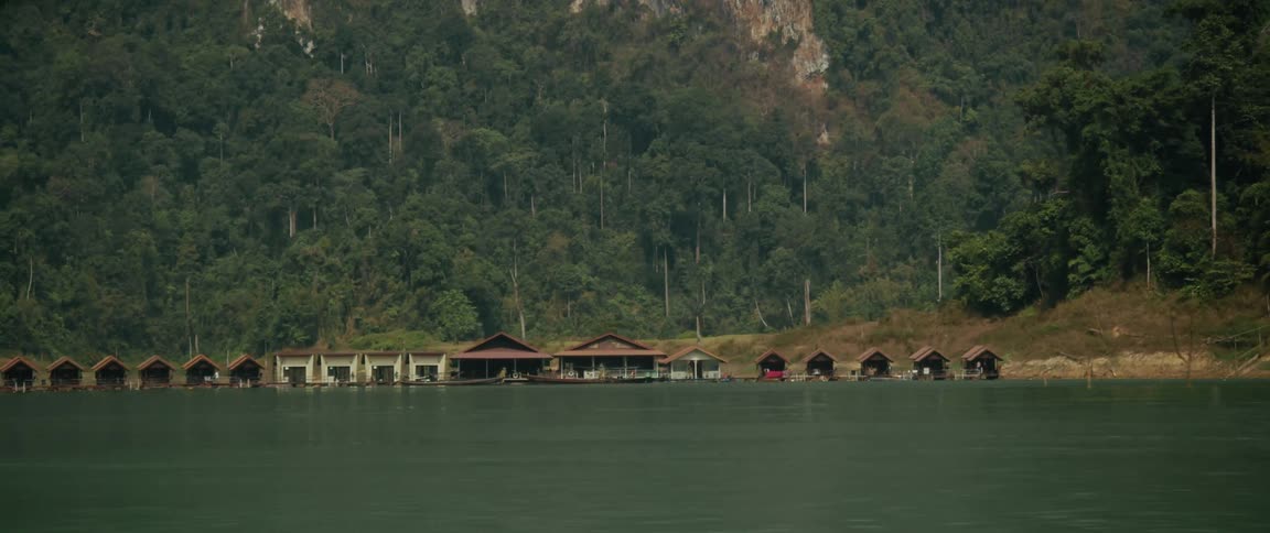 Floating bungalows sit on a tranquil green lake at the base of a lush jungle-covered mountain