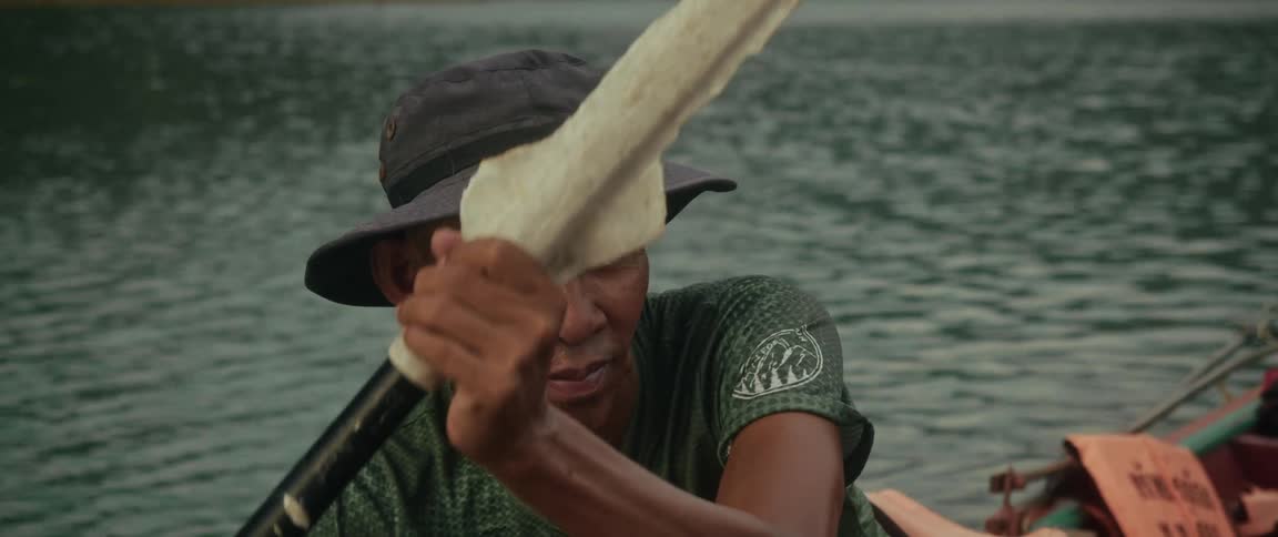 A focused Asian man wearing a bucket hat paddles his boat across calm waters in slow motion