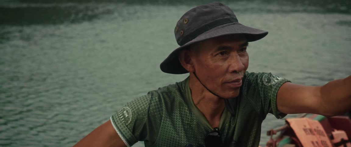 A close-up shot captures the thoughtful expression of an older Asian man in a boonie hat on a moving boat