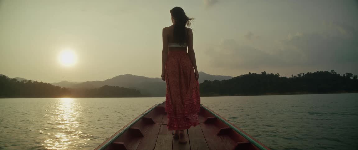 A serene cinemagraph shows a woman in a long flowing skirt standing on the bow of a boat enjoying the beautiful sunset over a mountain lake