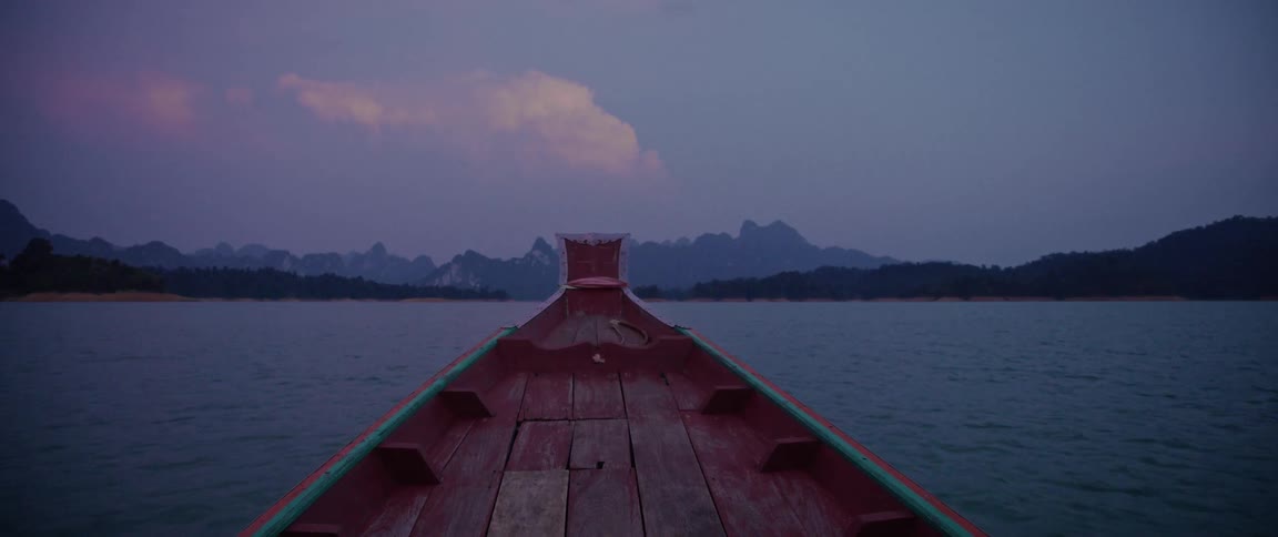 A serene point-of-view from a traditional wooden long-tail boat gliding across a lake towards mountains at dusk