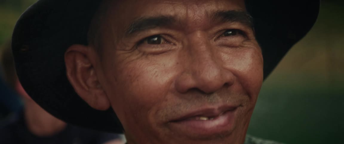 A warm, close-up portrait of an elderly Asian man in a hat, whose serious expression slowly breaks into a genuine, kind smile