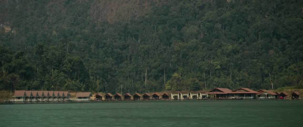 A scenic view of floating bungalows on a calm lake at the base of a lush, jungle-covered mountain