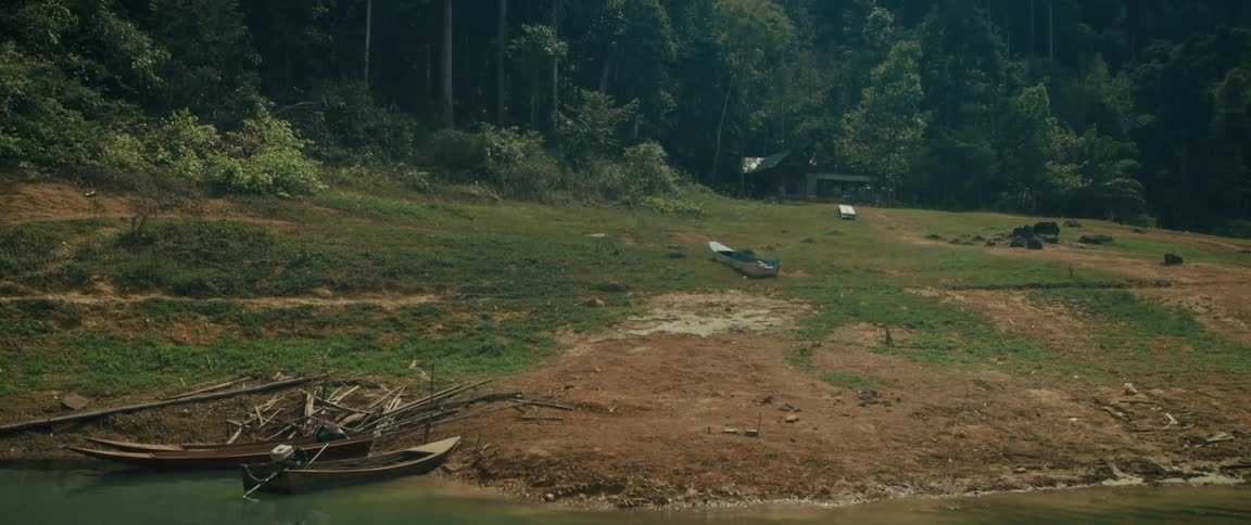Longboats are moored on the muddy bank of a remote jungle river, with a small shack nestled in the lush green forest