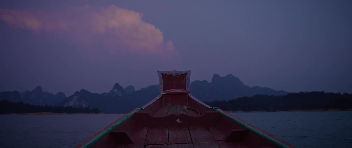 A point-of-view shot from the bow of a wooden boat cruising across a calm lake toward majestic mountains at dusk