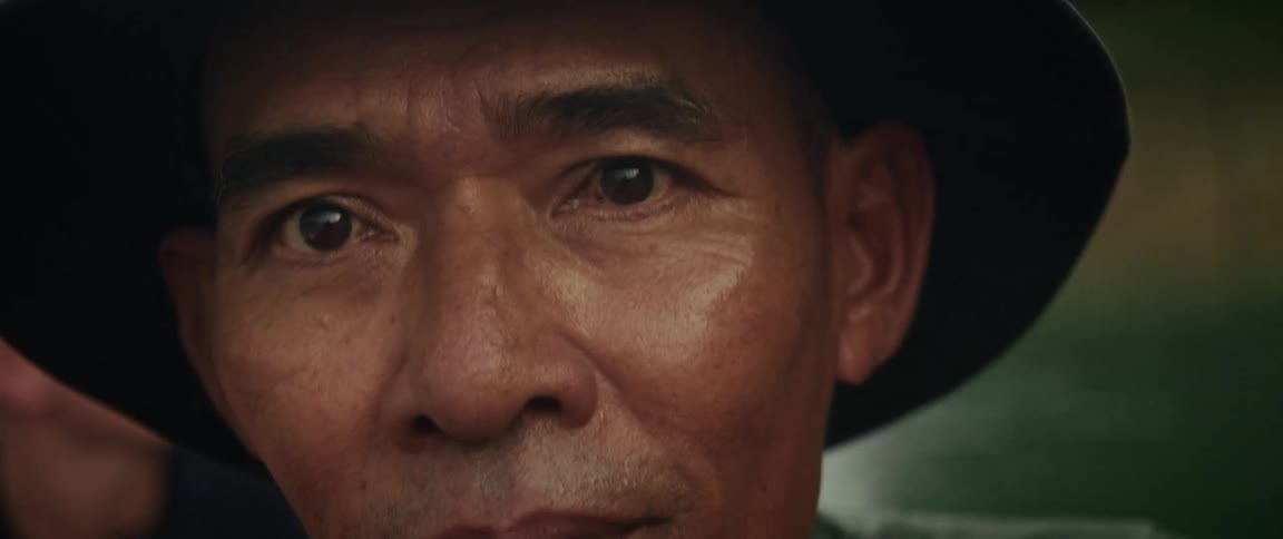 An intimate close-up of a weathered Asian man in a bucket hat turning his head to look thoughtfully at the camera