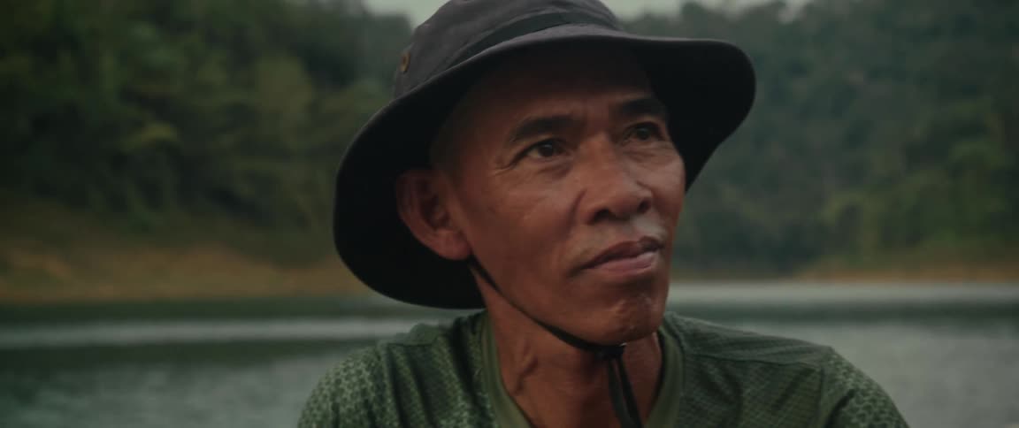 A close-up of a thoughtful elderly Asian man in a hat as he paddles a boat across a tranquil lake in slow motion
