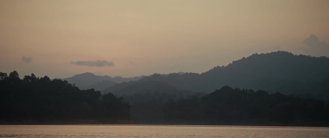 A peaceful, atmospheric shot of layered mountain silhouettes across a tranquil lake during a warm sunset