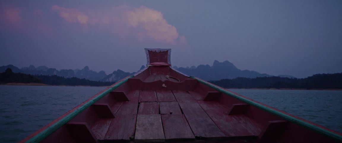 A serene journey on a traditional long-tail boat across a lake towards majestic mountains during a moody twilight