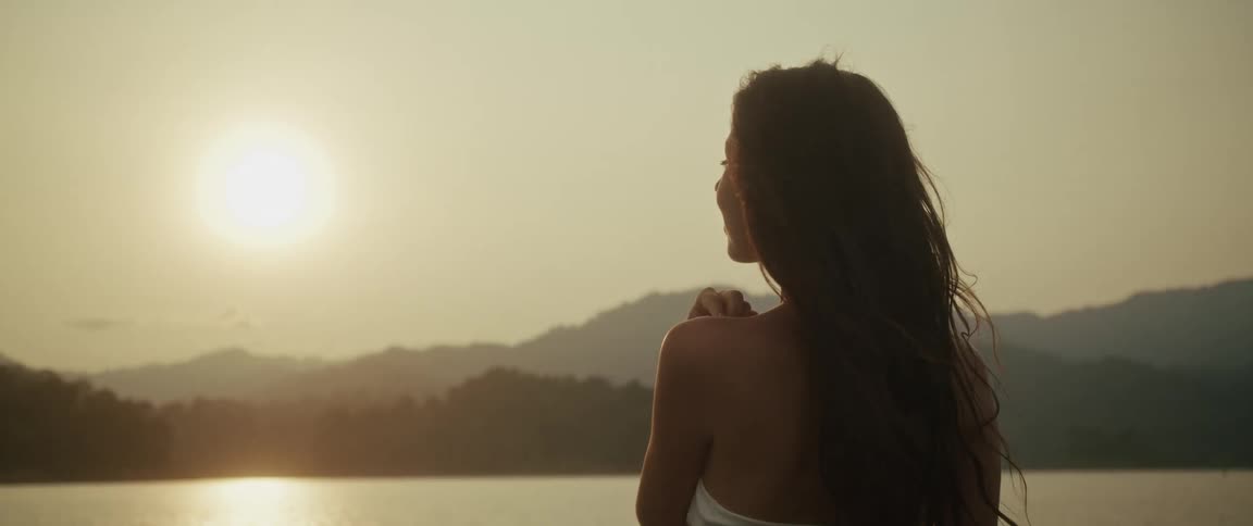 A woman with long flowing hair watches the golden sunset over a mountain lake before turning to smile at the camera in slow motion