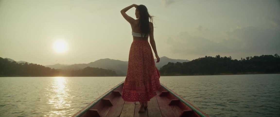 A beautiful woman in a flowing skirt stands on the front of a boat, enjoying a serene sunset over a mountain lake