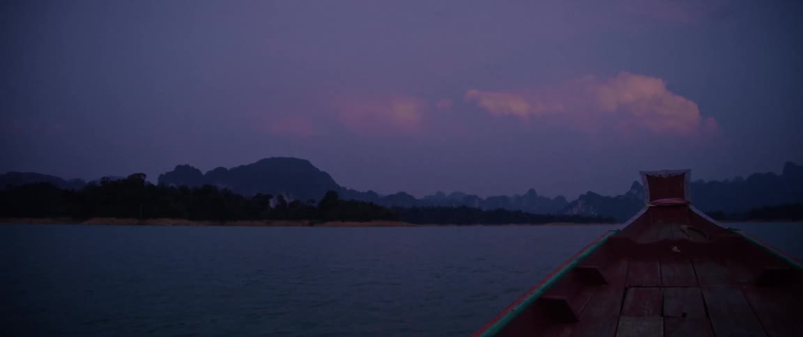Point-of-view from a traditional wooden boat sailing across a serene lake at twilight, revealing a mountainous landscape under a purple sky