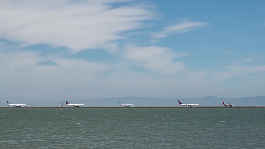A time-lapse of a busy airport runway next to the water shows a continuous stream of airplanes taking off and landing