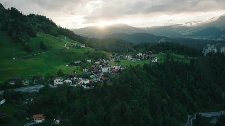 A slow aerial pullback reveals a quaint European village tucked into the mountainside as the sun sets over the peaks