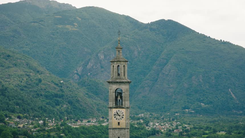 The bell of an old stone clock tower chimes in front of a lush green mountain landscape