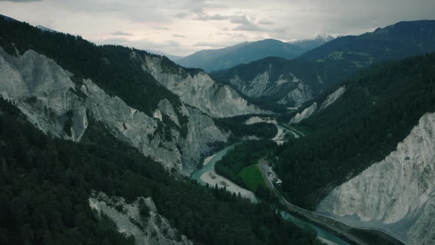 A majestic aerial flight over a dramatic mountain gorge reveals a turquoise river winding through a vast, forested valley under an overcast sky