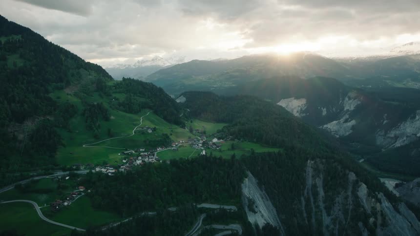 A dramatic aerial view captures a picturesque mountain village nestled in a lush valley as the sun sets over the Swiss Alps