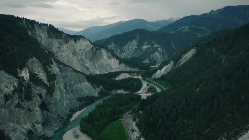 A majestic aerial shot follows a red train as it journeys through a dramatic, forested mountain canyon alongside a winding turquoise river