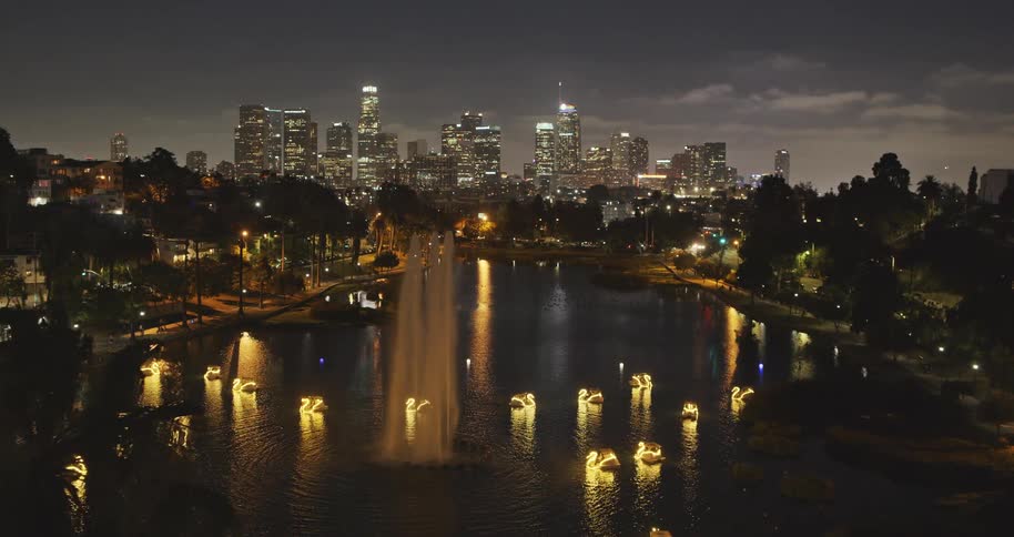 A beautiful nighttime aerial shot of Echo Park Lake, featuring a glowing fountain, lit-up swan boats, and the dazzling Downtown Los Angeles skyline