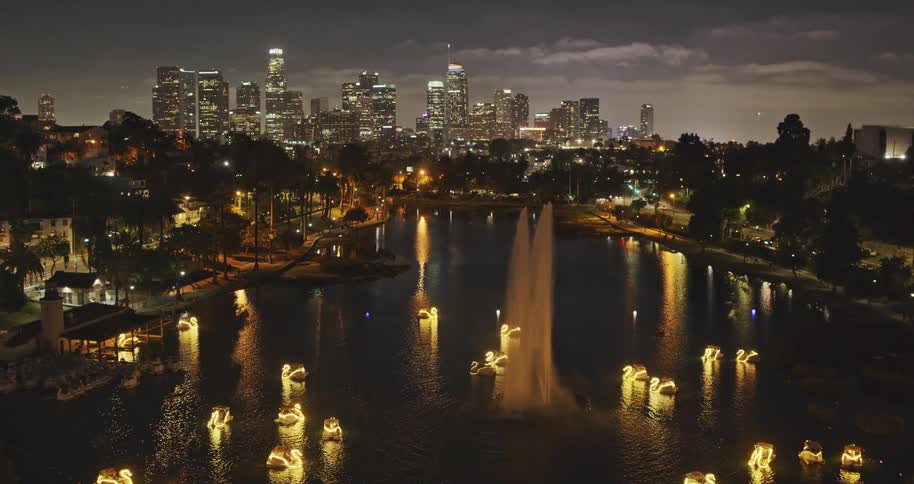 A stunning nighttime view of the Los Angeles skyline over Echo Park Lake, featuring a brilliant fountain and glowing swan boats